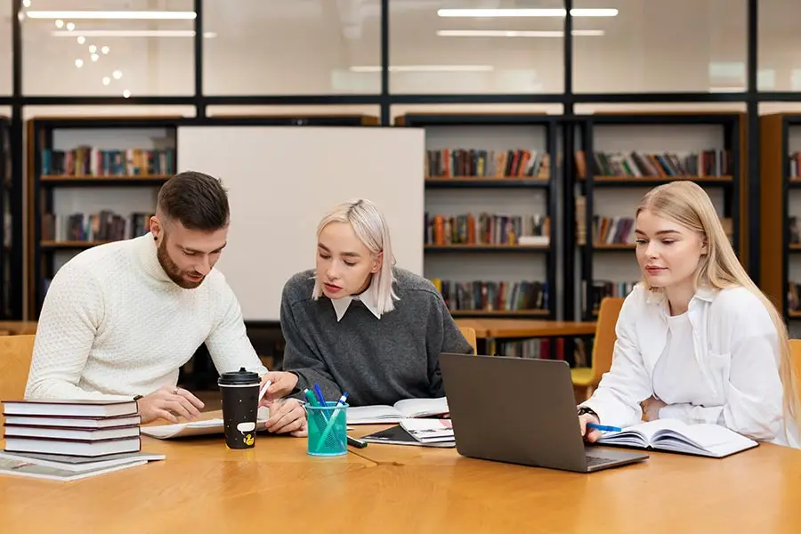 Three young adults studying together in a library with books and a laptop.